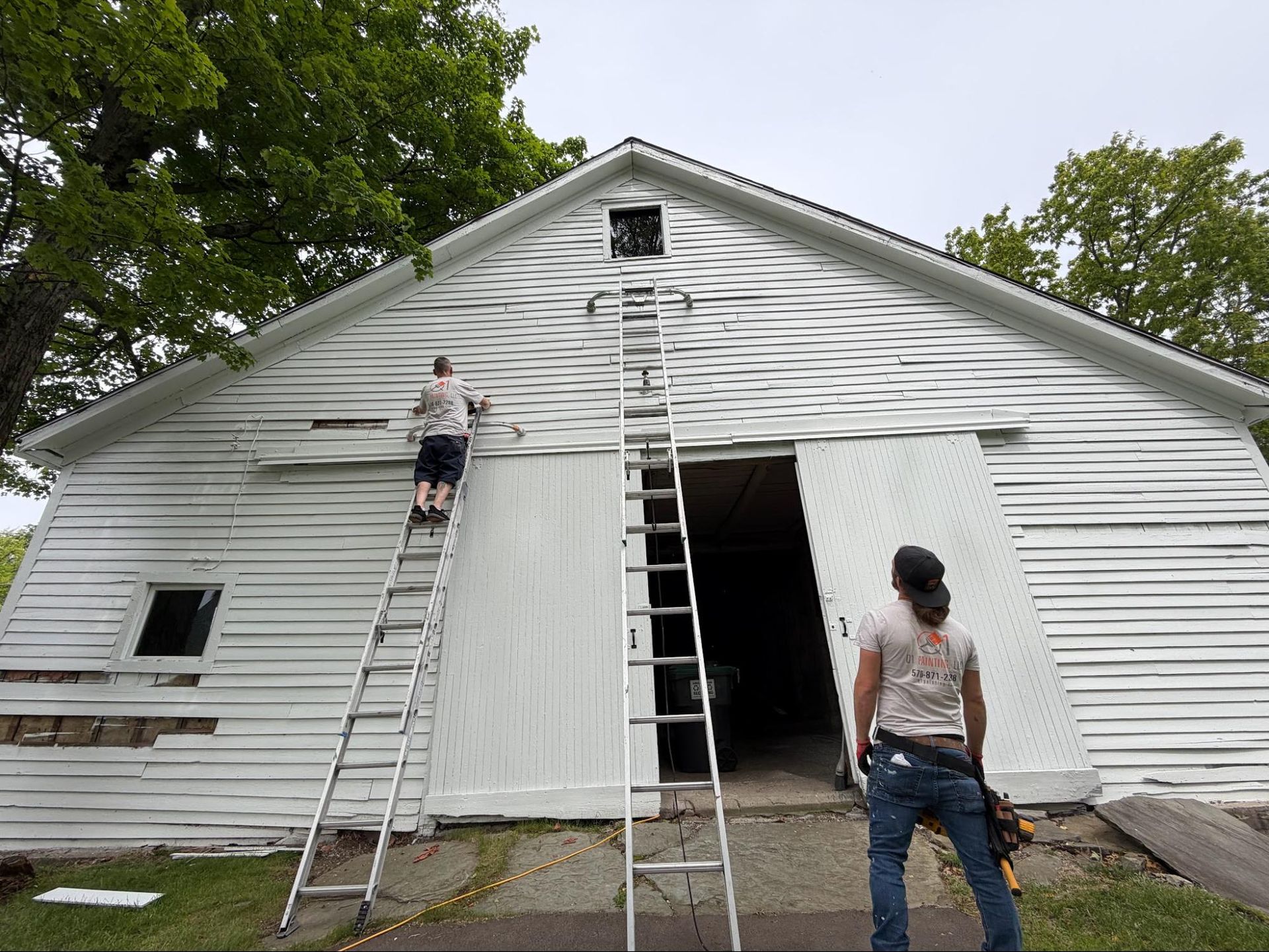 Workers on ladders painting a white barn with open doors outdoors