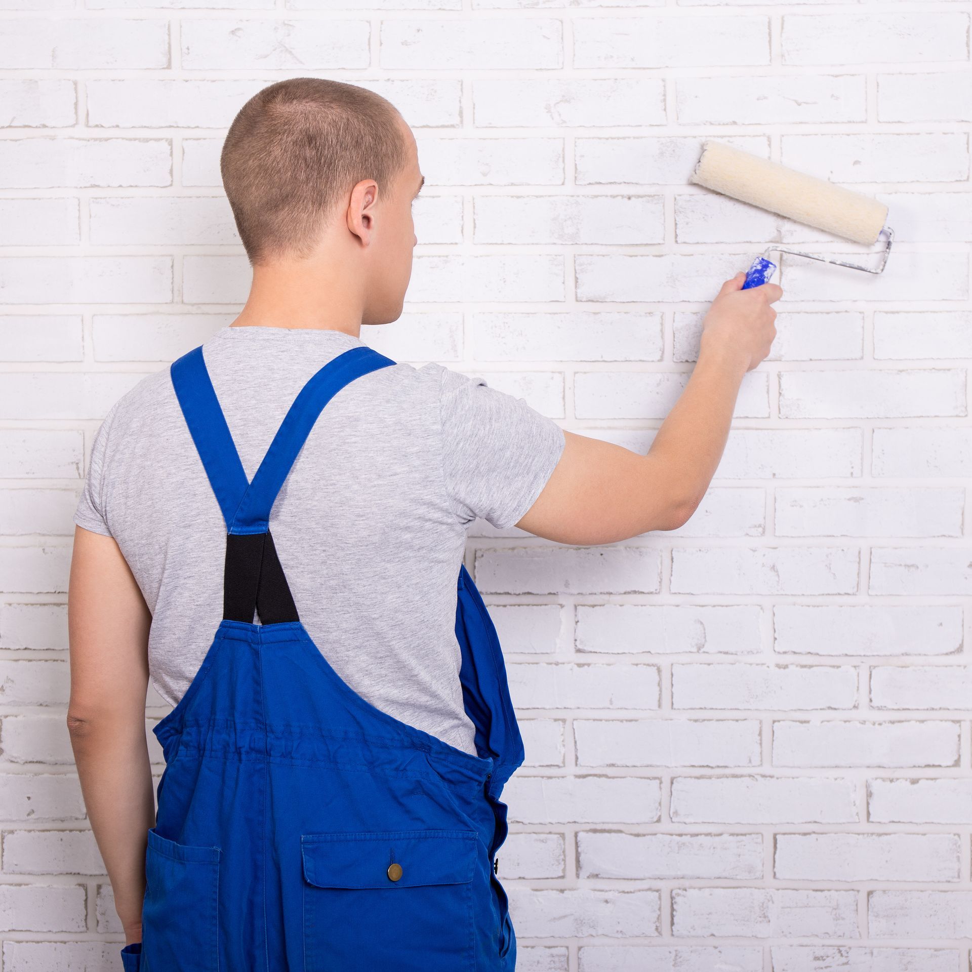 A man in blue overalls is painting a white brick wall with a paint roller.
