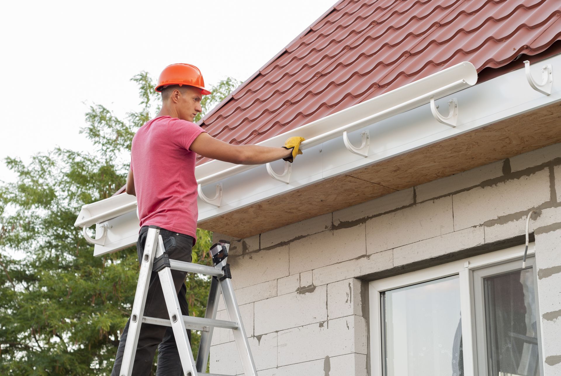 Person on ladder cleaning gutters on a house; wearing orange hard hat and red shirt.