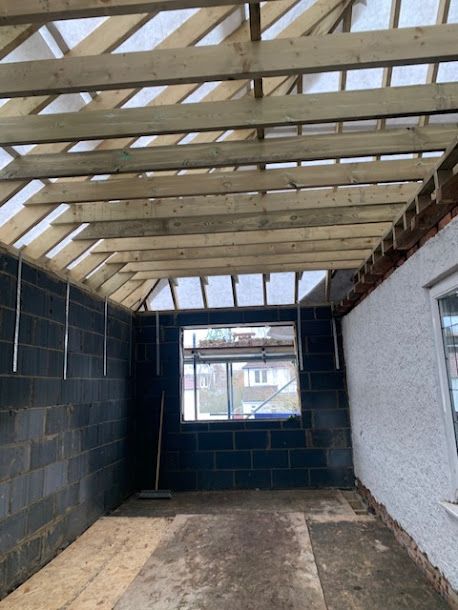Interior of a partially built room with exposed roof beams, cinderblock walls, and a window opening.
