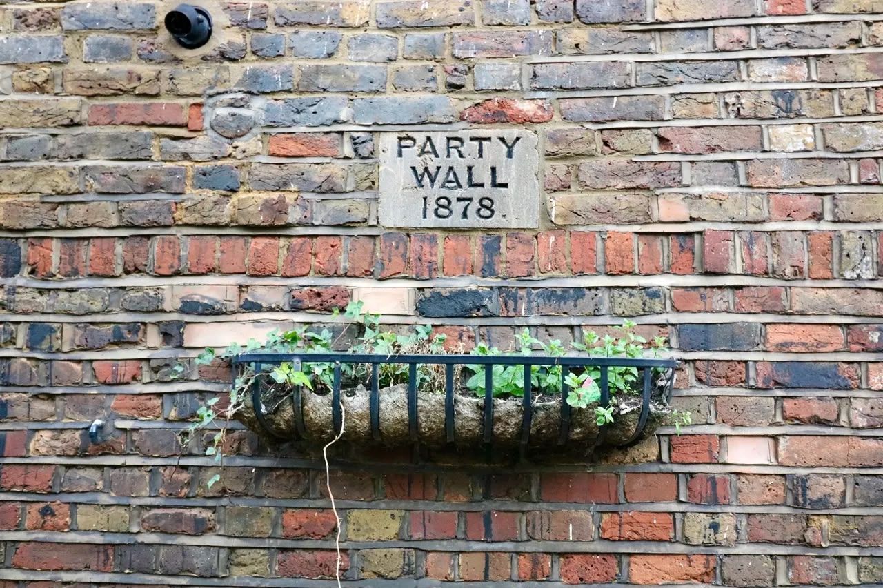 Brick wall plaque reading “Party Wall 1878” with a small flower planter beneath it