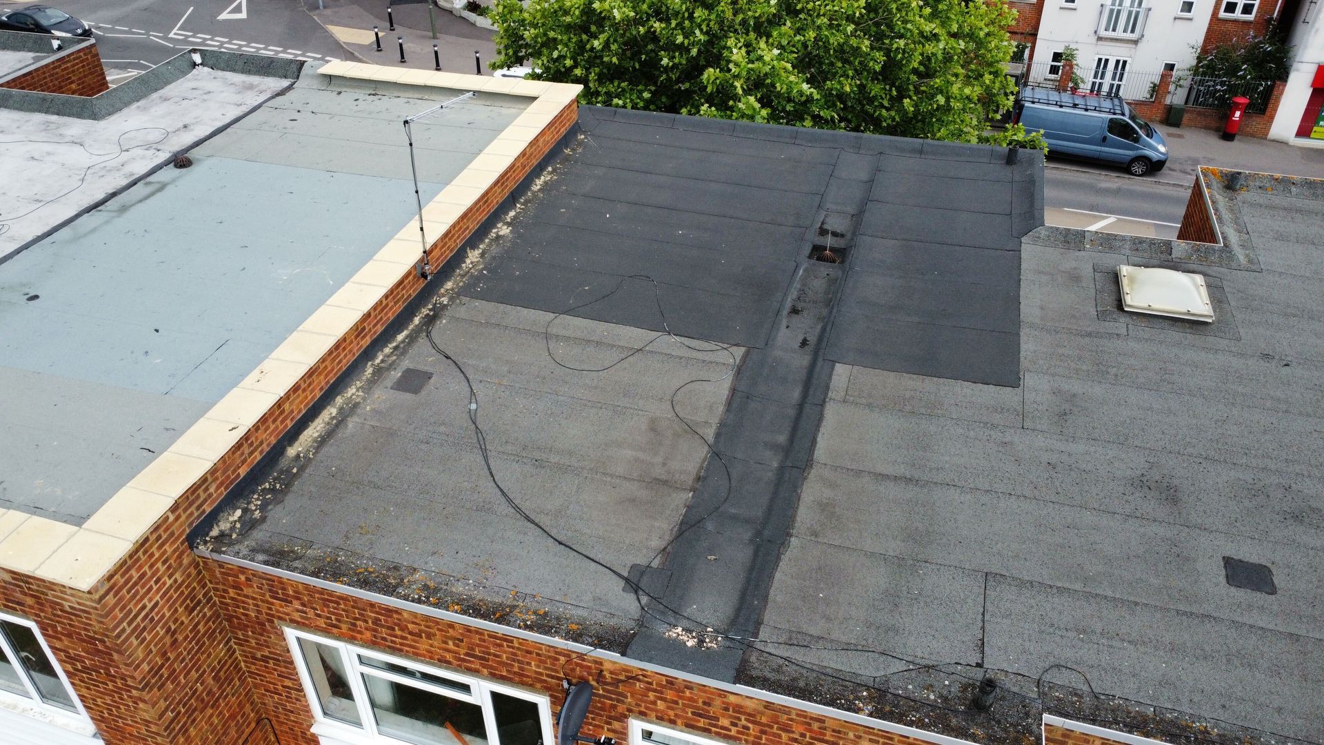 Rooftop with patched flat roofing, vents, and brick walls beside a tree-lined street below