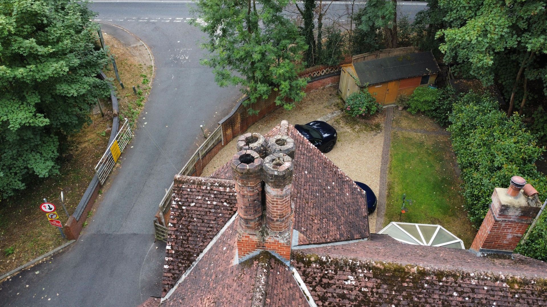 Rainy view over a winding road and tiled rooftops with chimneys, trees, and a parked car.