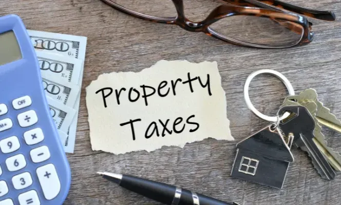 Property taxes note beside calculator, glasses, keys, house keychain, and cash on a desk