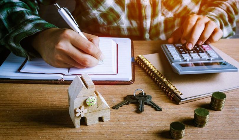 Hands writing in a notebook beside a calculator, house model, and coins on a wooden desk