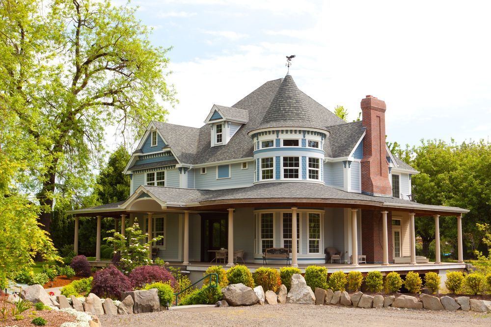 Large Victorian-style house with wraparound porch, gray roof, and colorful landscaping on a sunny day