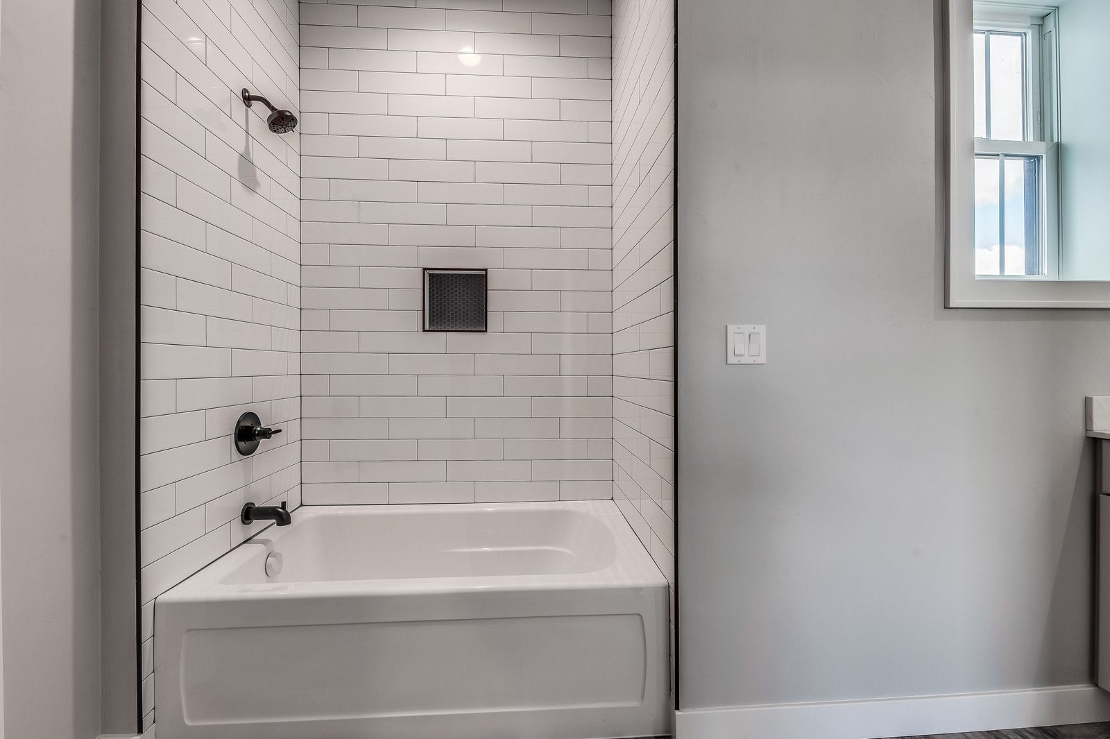 White tiled bathtub and shower alcove beside a gray bathroom wall with a small window.