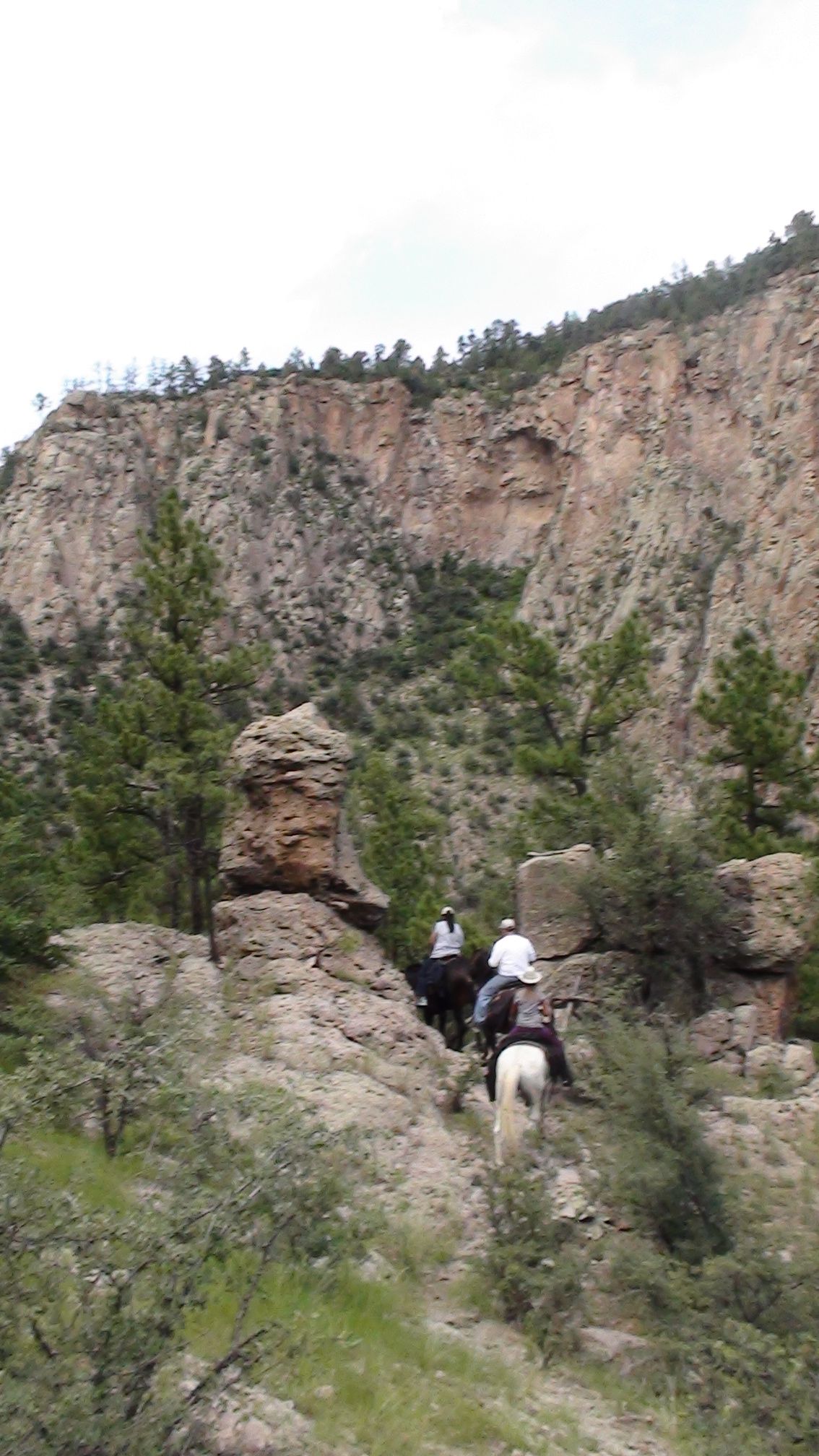Two horseback riders trail through a rocky canyon with steep cliffs and sparse greenery.
