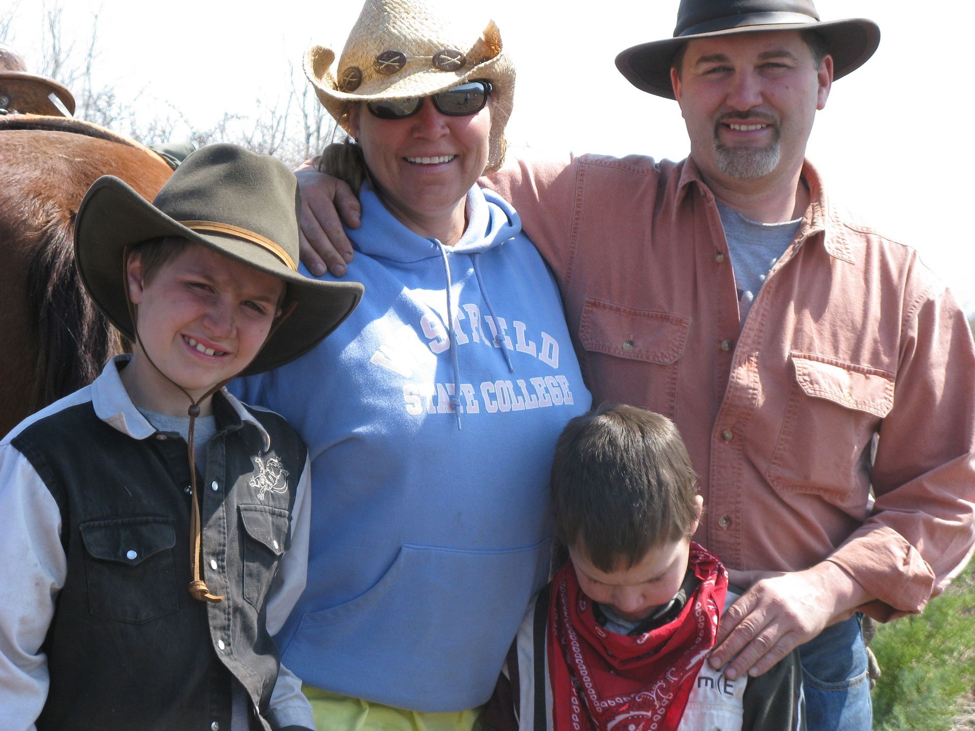 Four people in cowboy hats smiling outdoors, with a child in the foreground wearing a red scarf.