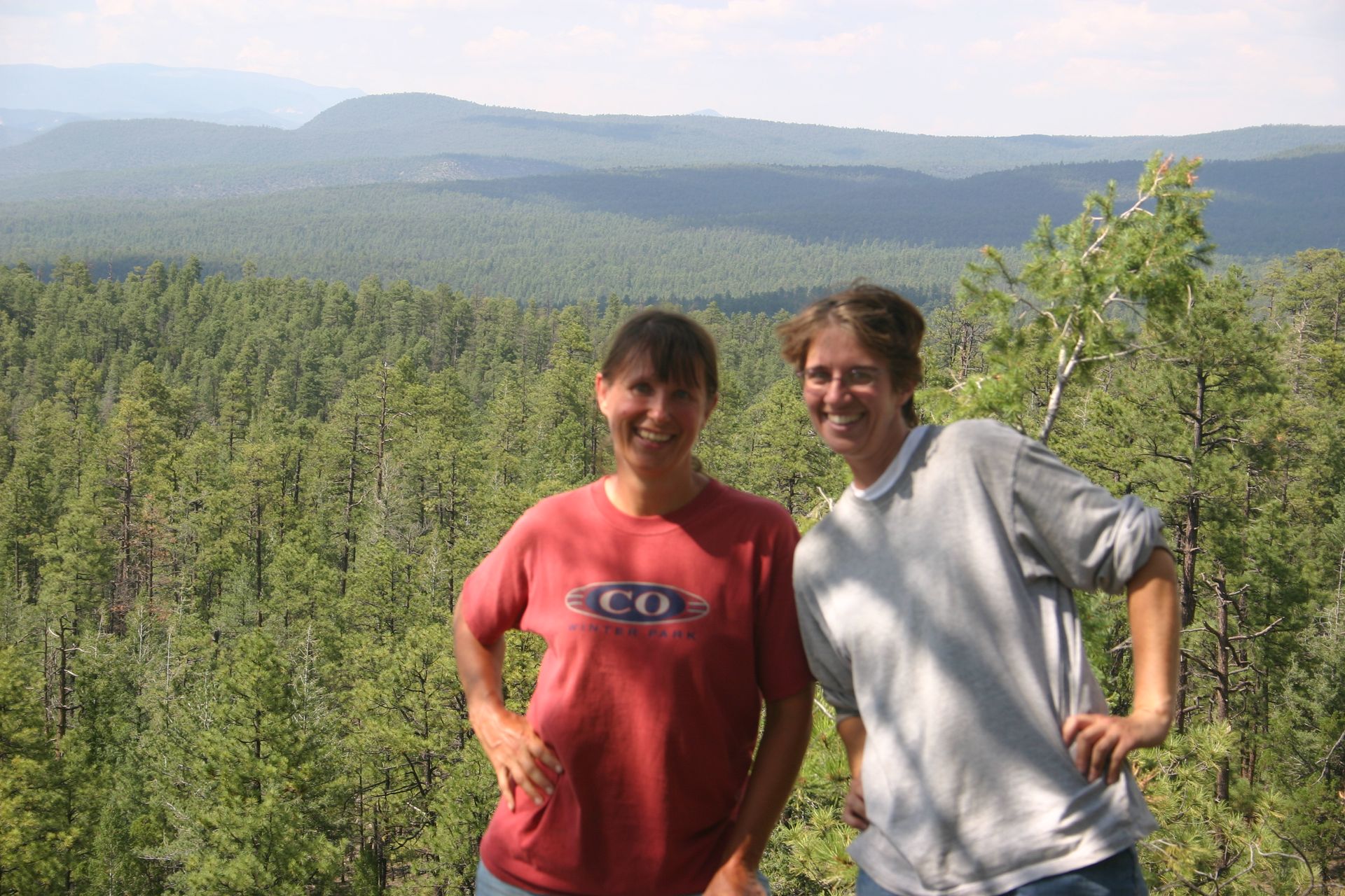 Two hikers standing on a mountain overlook with forested hills in the background