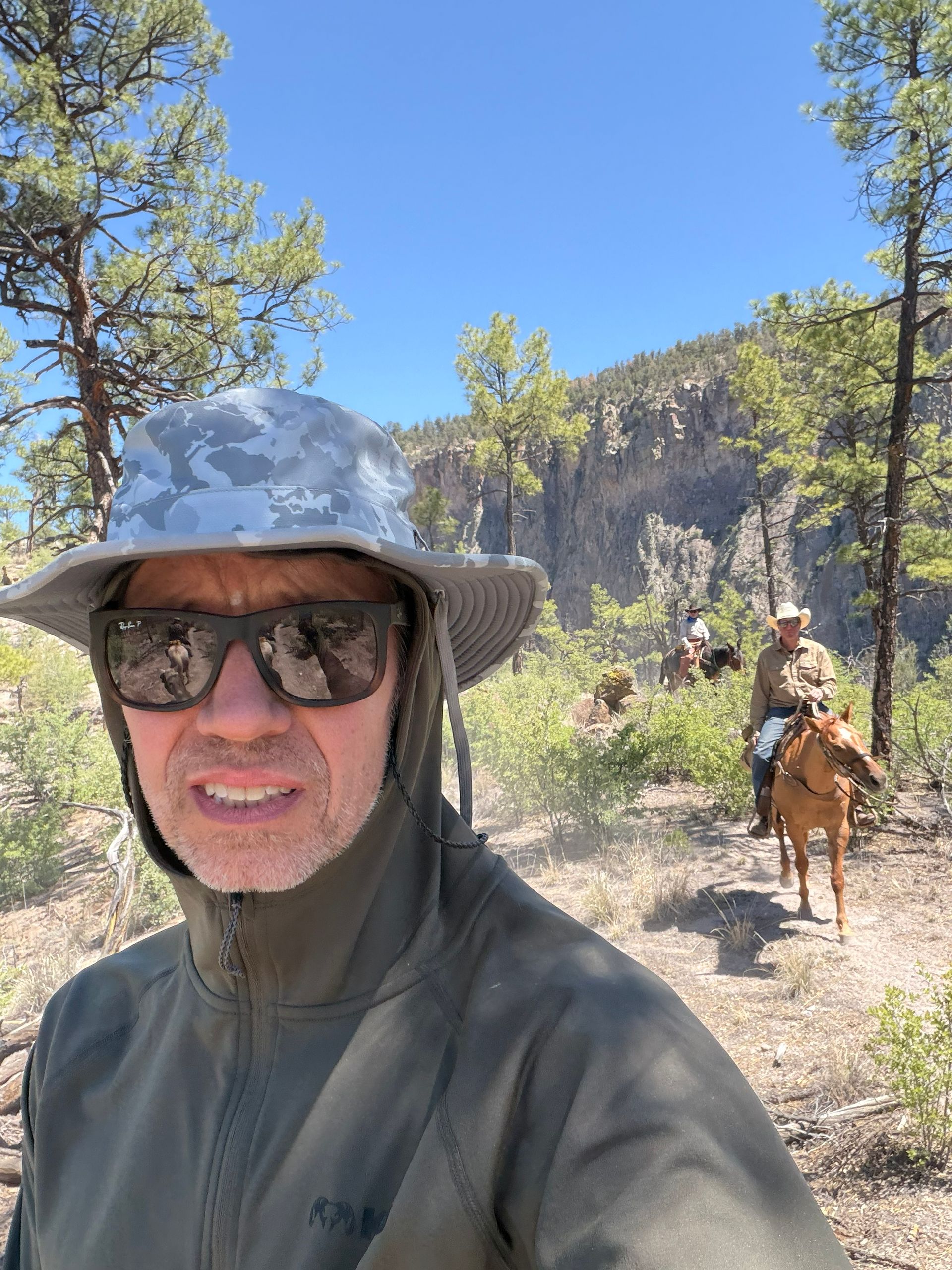 Man in sunglasses and gray hat on a sunny mountain trail with a horse and rider behind him