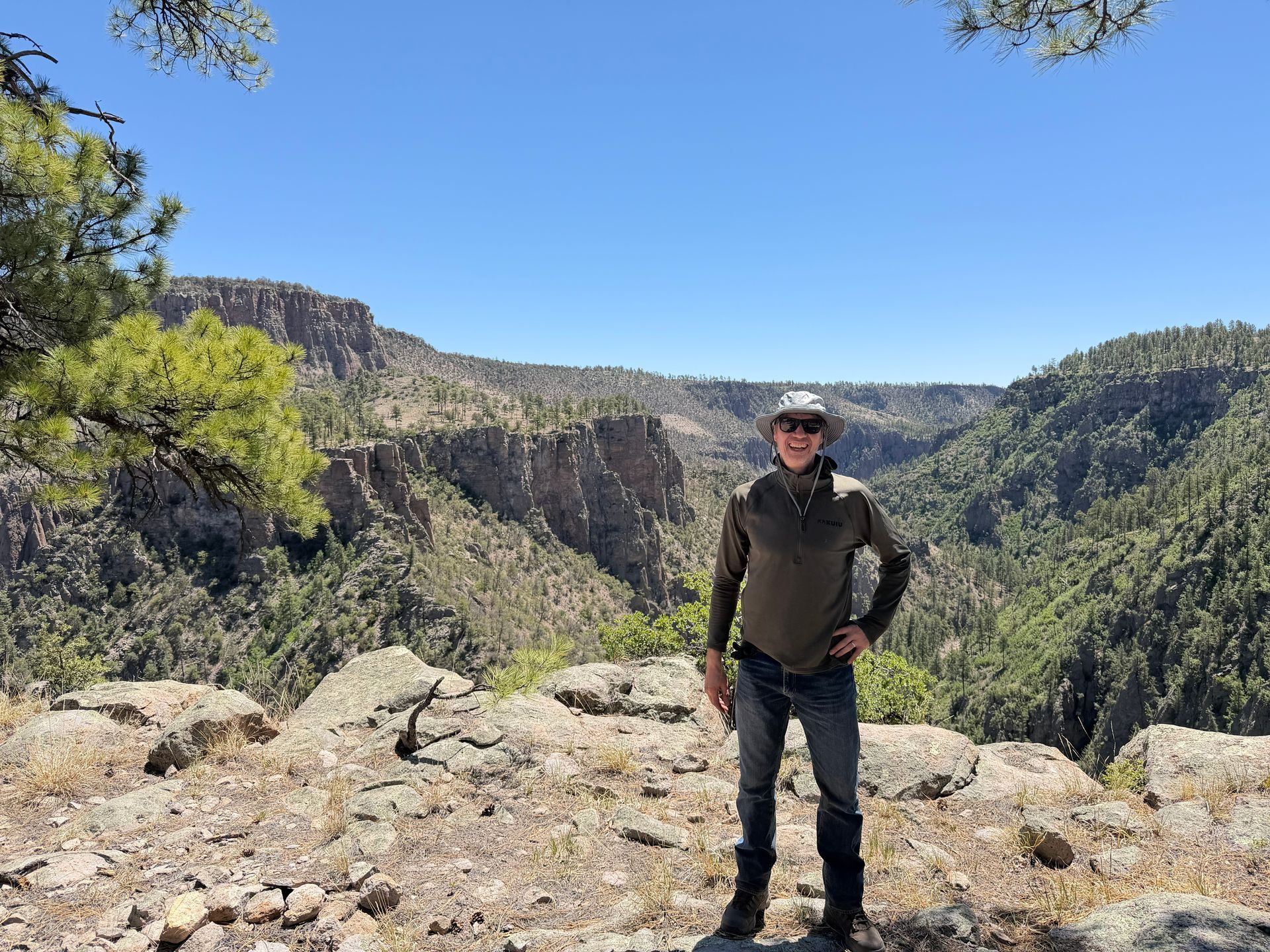 Person standing on a rocky overlook in a canyon, with green cliffs and blue sky behind.