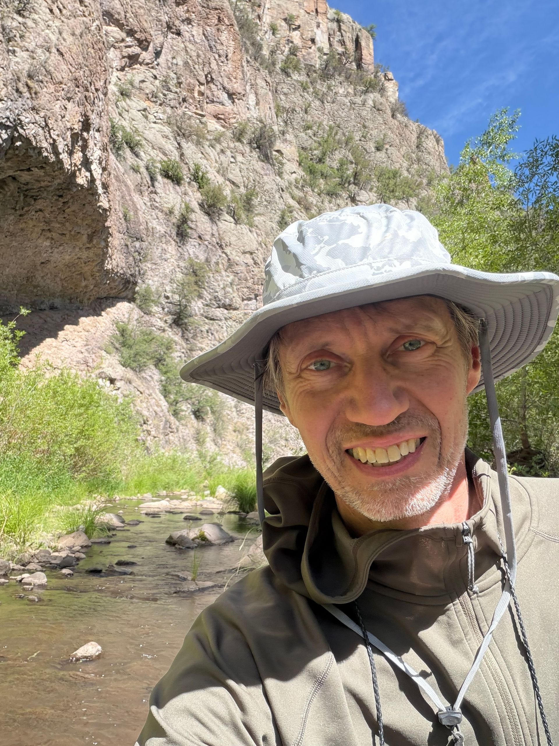 Smiling hiker in a gray sun hat by a rocky stream and cliff under a blue sky