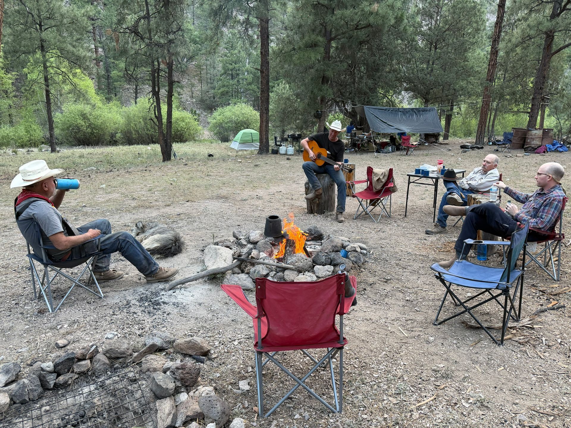 People relaxing around a campfire in folding chairs at a wooded campsite, with a tent in the background