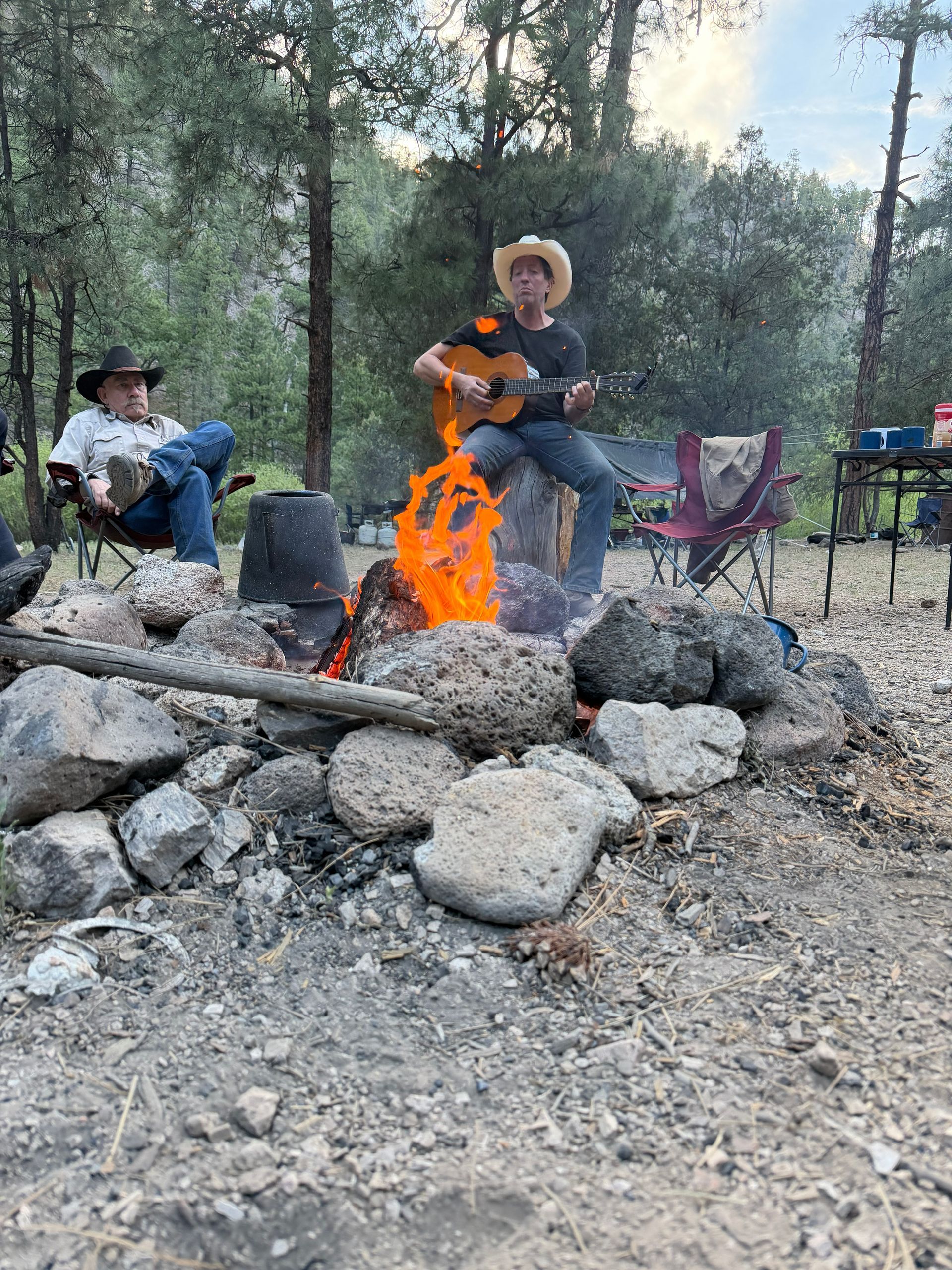 People gathered around a campfire in a wooded area, with one playing guitar and others seated nearby.