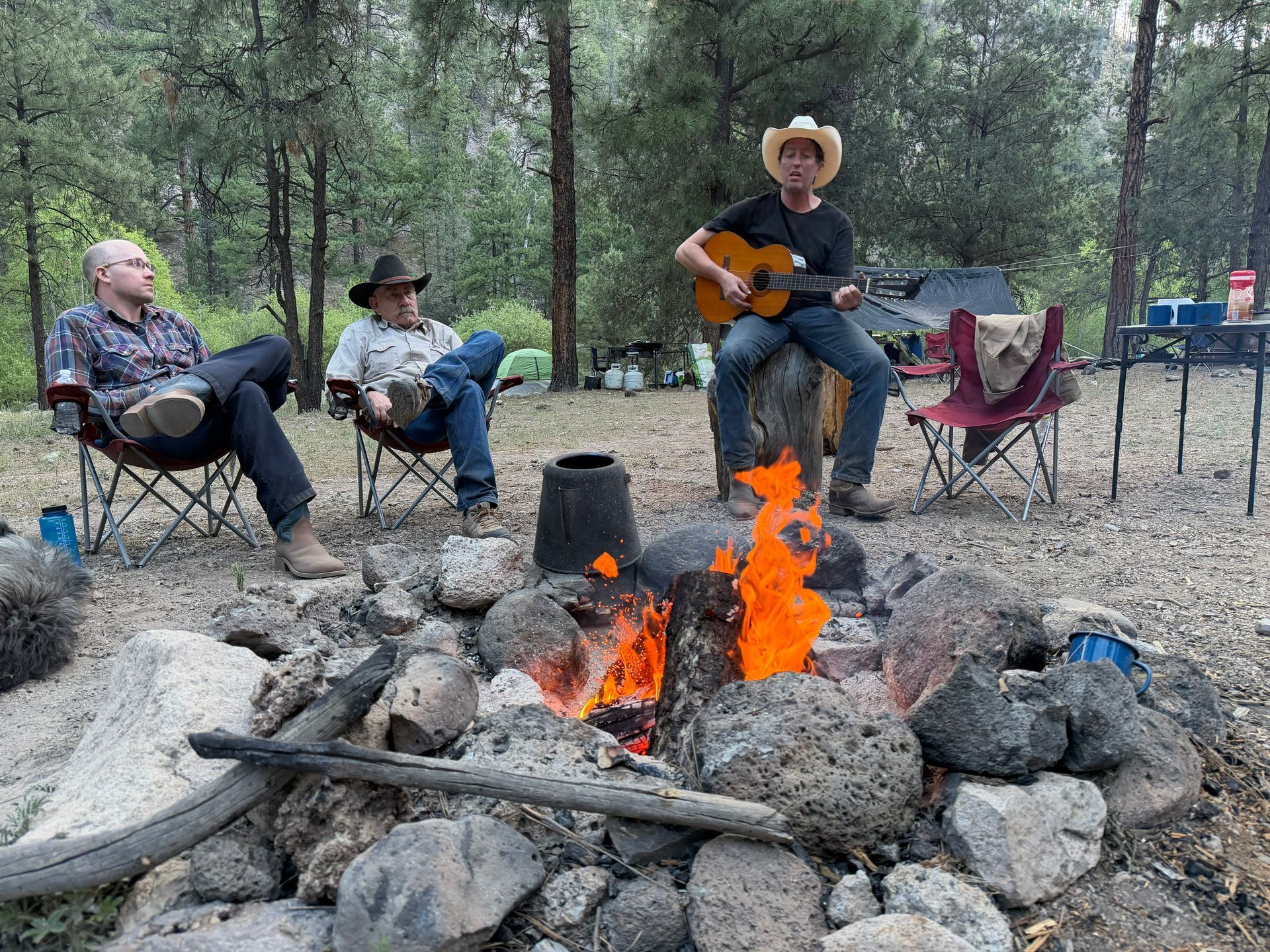People sitting in camp chairs around a campfire in a wooded campsite, one playing guitar.