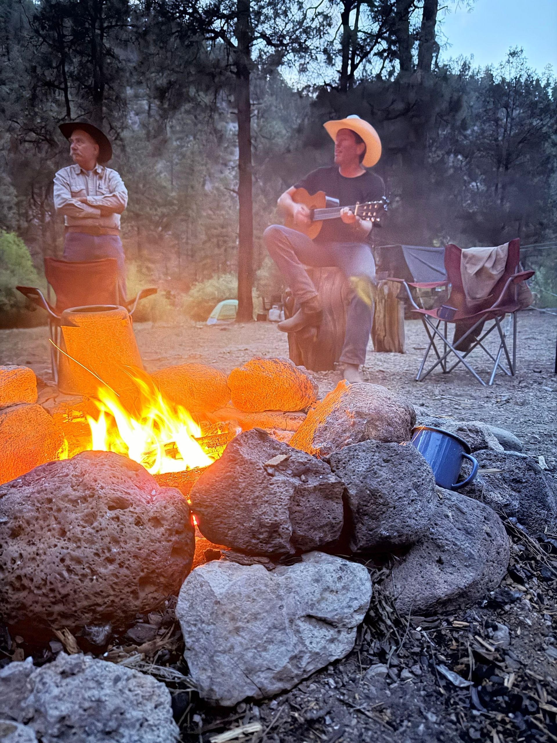 Two people sit by a campfire among rocks in a wooded outdoor setting.