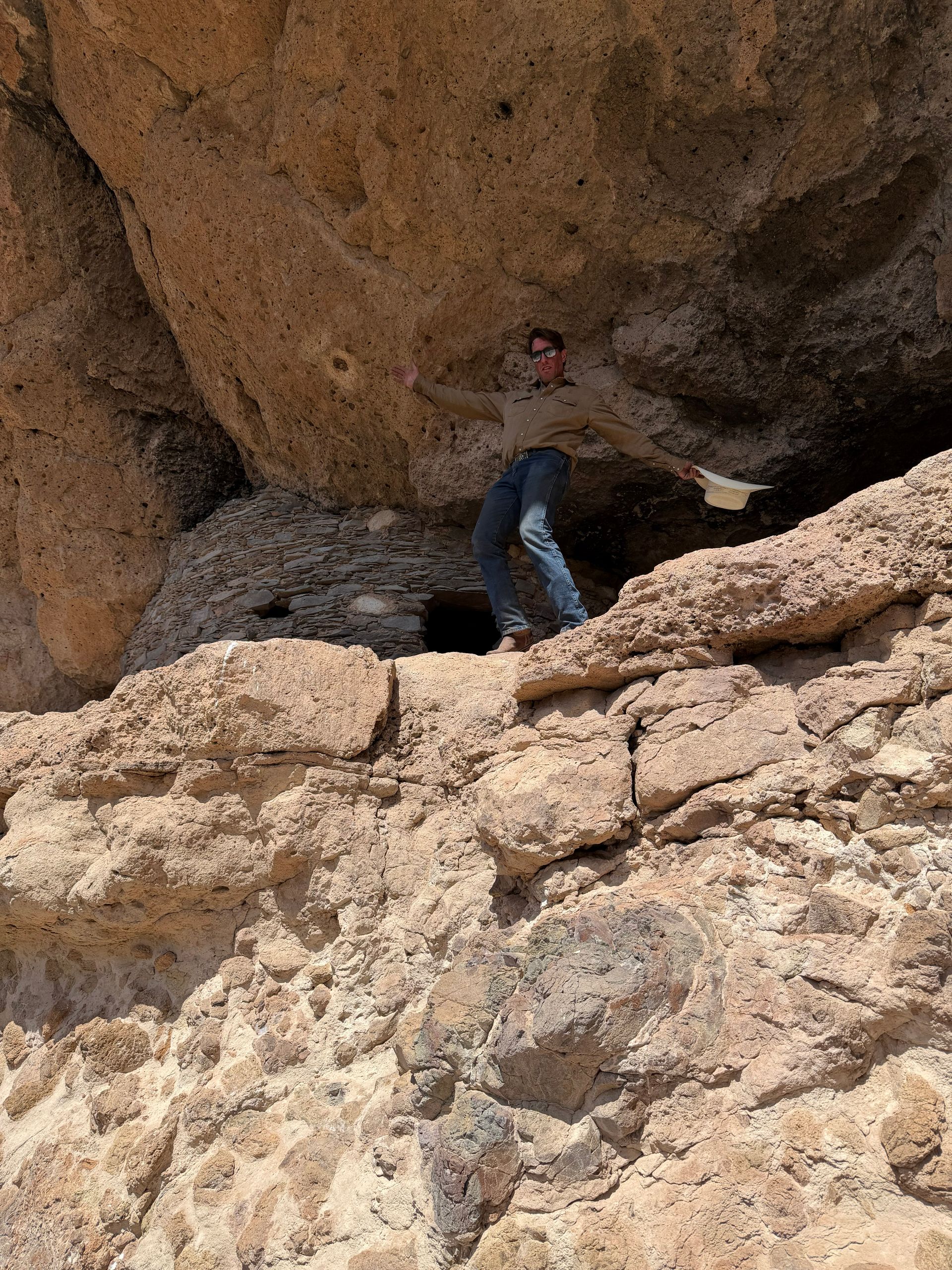 Person climbing under a rocky overhang in a desert cave, wearing blue jeans and a dark jacket.