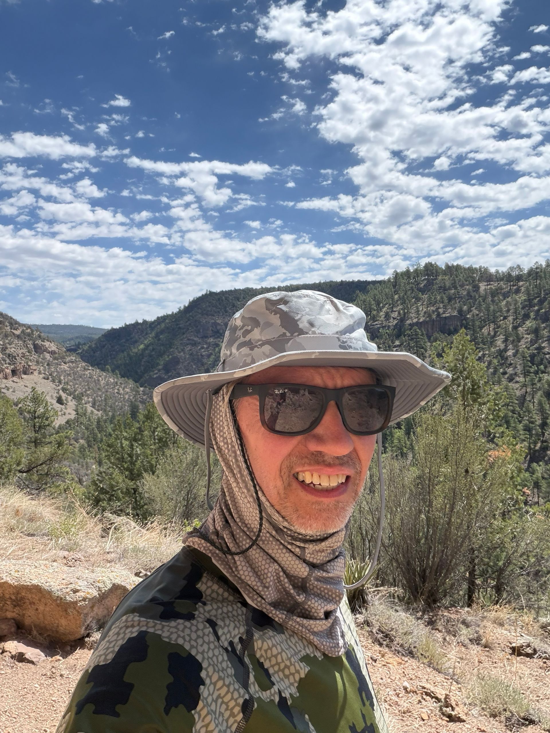 Smiling hiker in sun hat and sunglasses on a mountain trail, with rocky hills and blue sky behind.