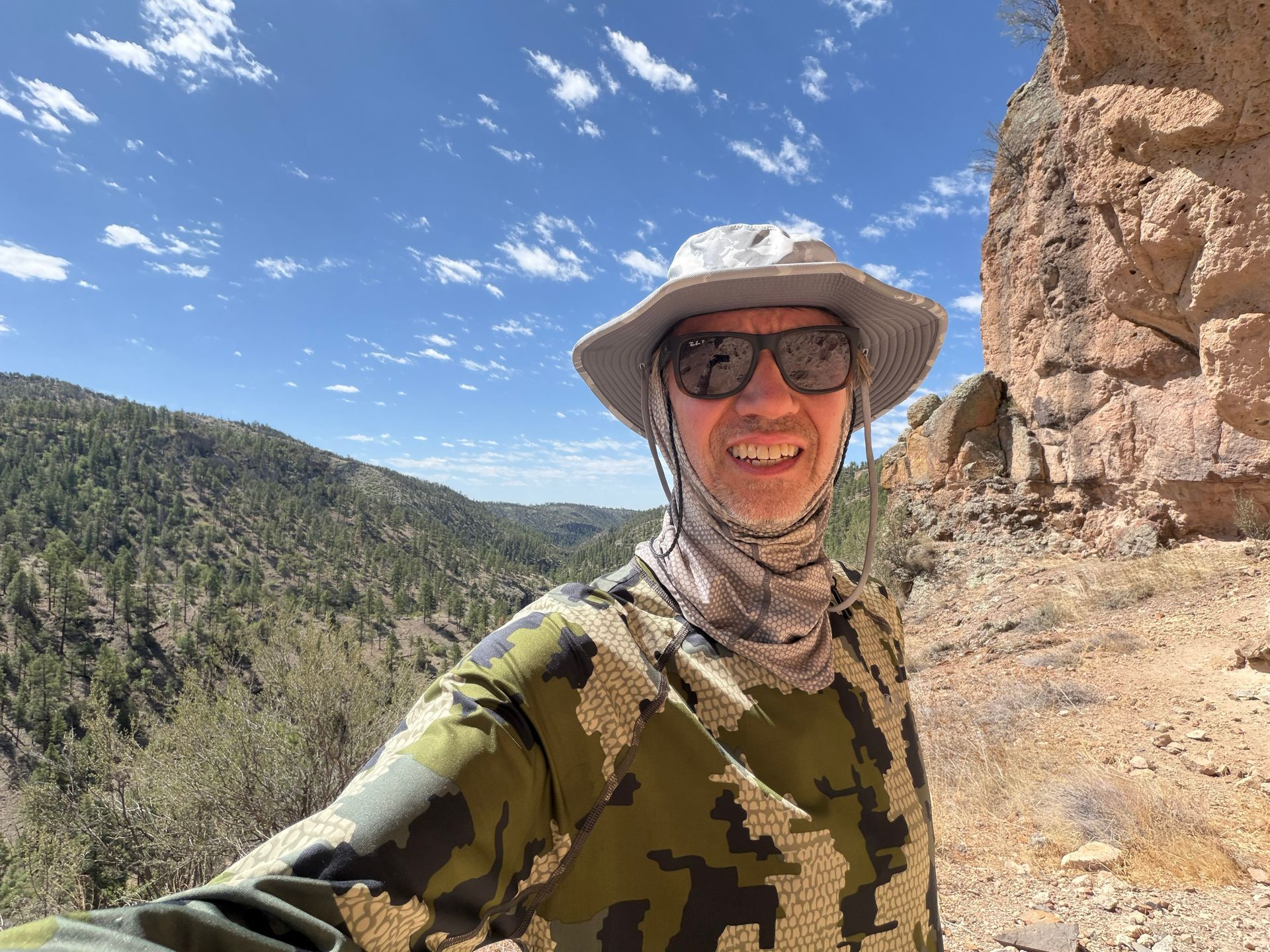 Smiling hiker in sun hat and sunglasses on a rocky cliff overlooking a forested canyon under a blue sky