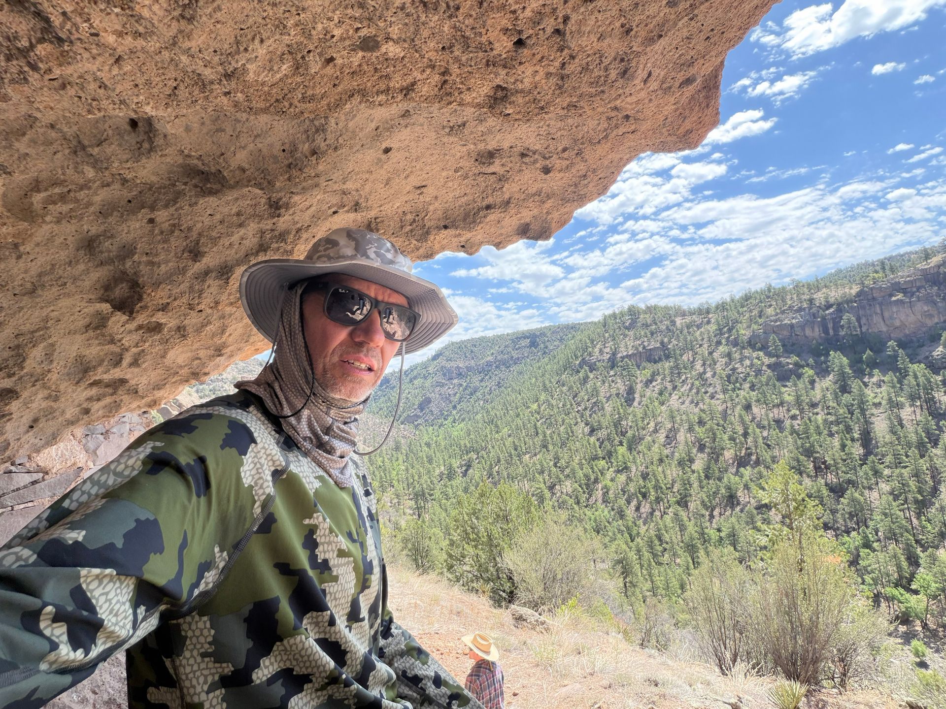 Person in camouflage and sunhat stands under a rock overhang overlooking a forested canyon.