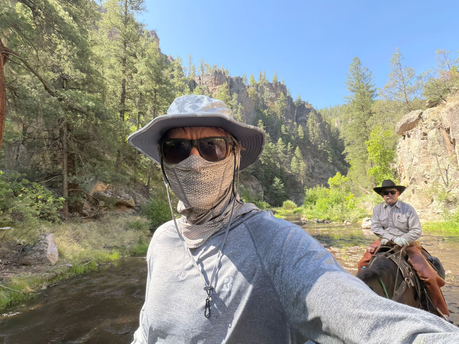 Person in sunhat and sunglasses standing by a river, with a rider on horseback in a wooded canyon.