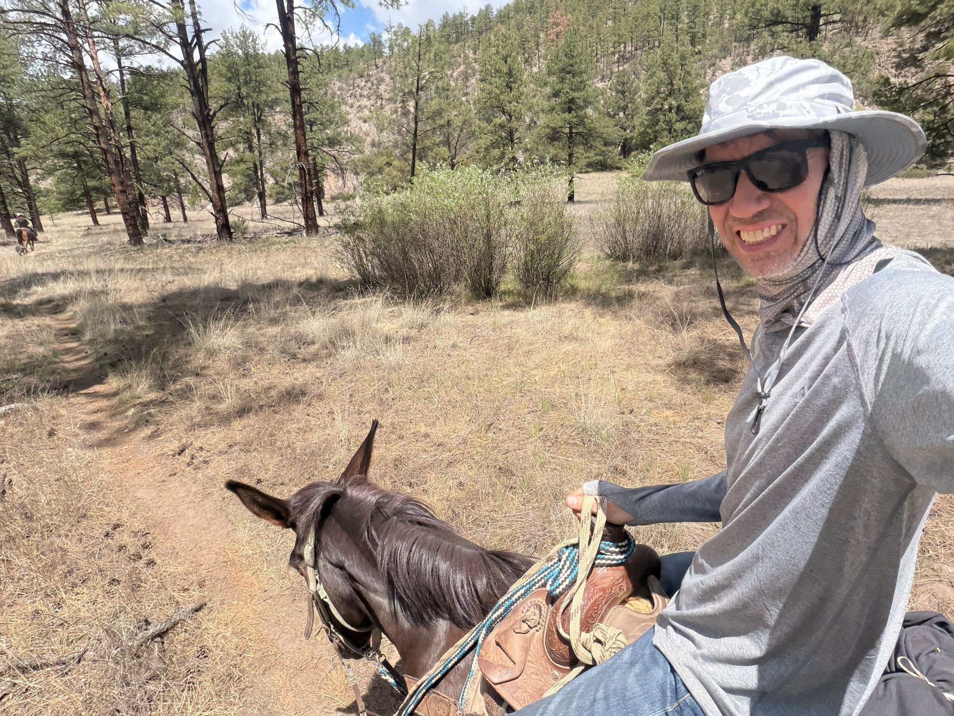 Person on horseback wearing sun hat and sunglasses, riding through a dry forest clearing.