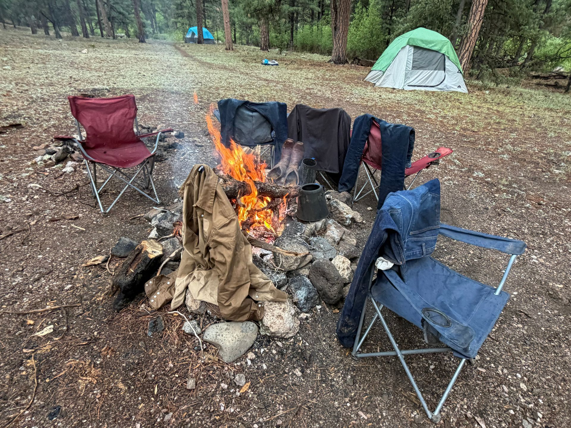 Campfire ring with several folding chairs in a wooded campsite, with tents in the background
