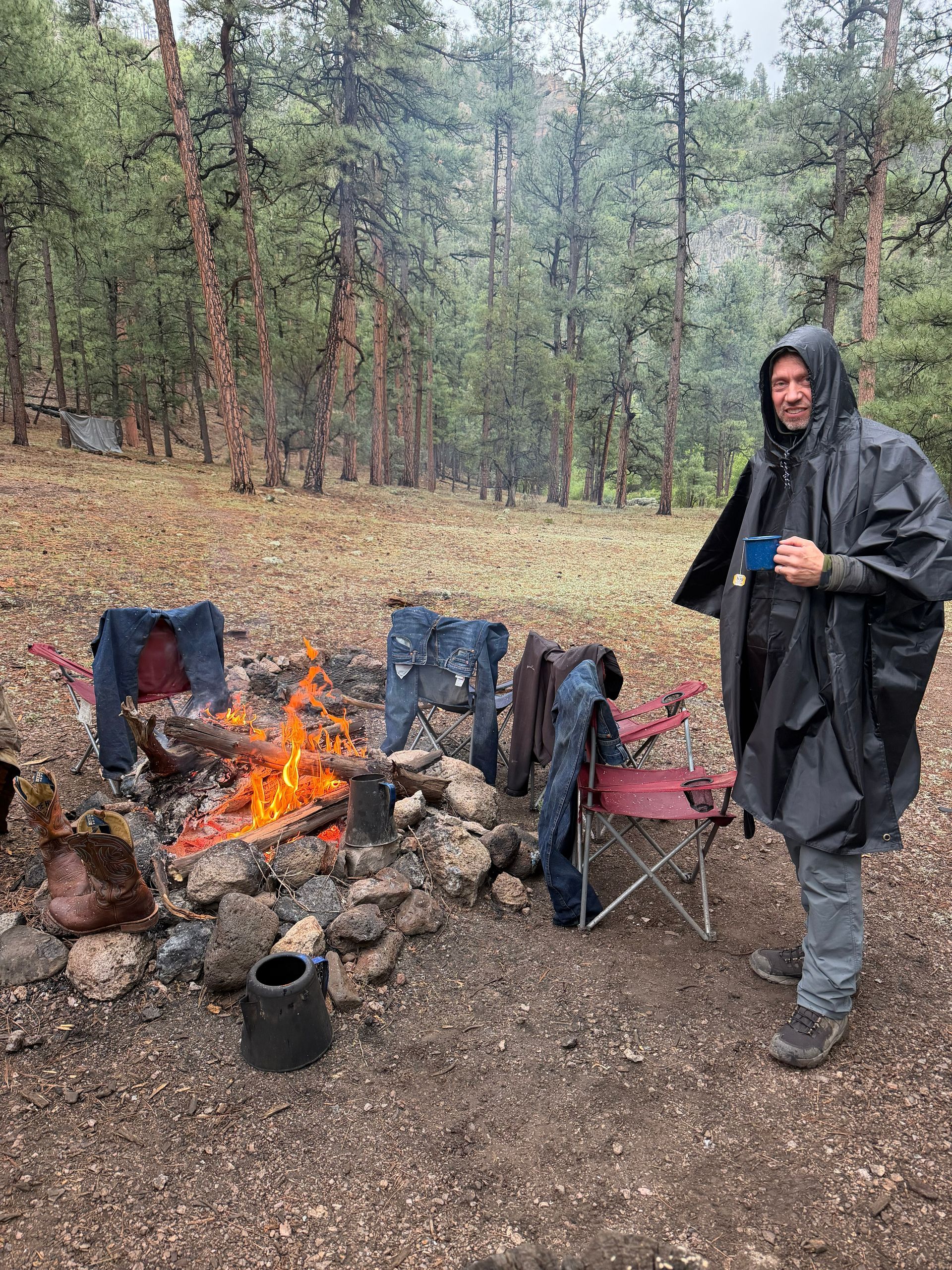 Person in a black poncho stands by a campfire in a wooded campsite with chairs and a kettle.