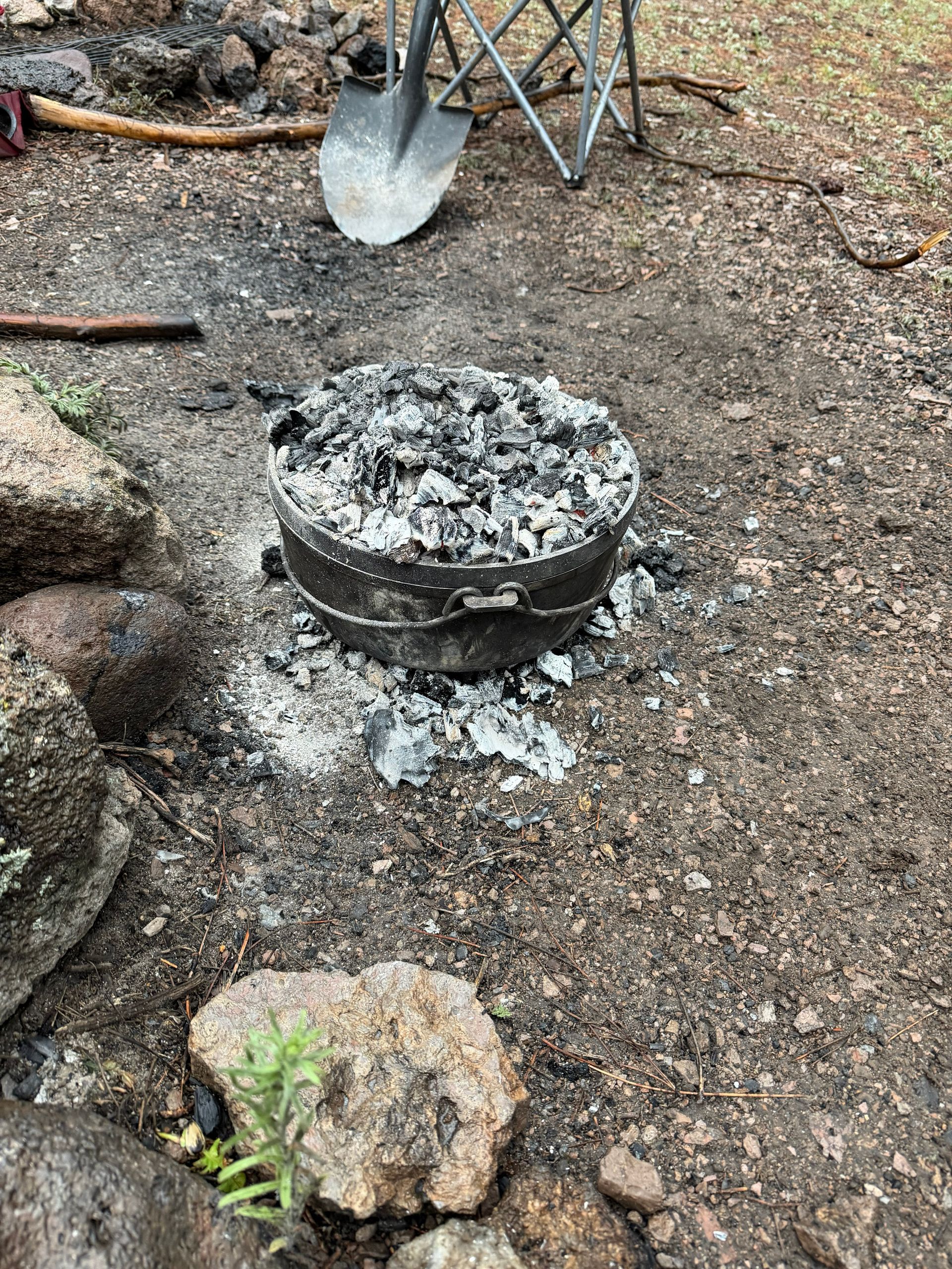Charcoal grill or fire pit filled with ash on rocky ground beside a shovel and folding chair