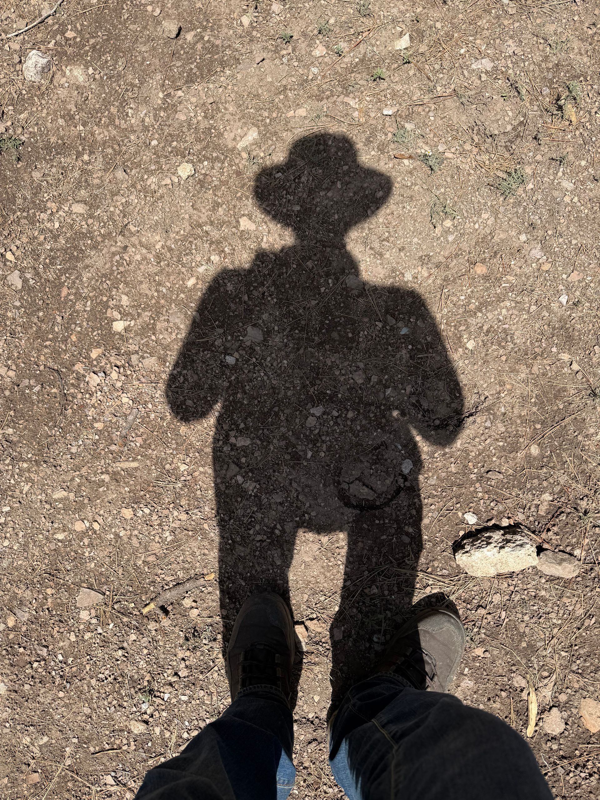 Shadow of a person wearing a hat on a gravel path, with shoes visible at the bottom.