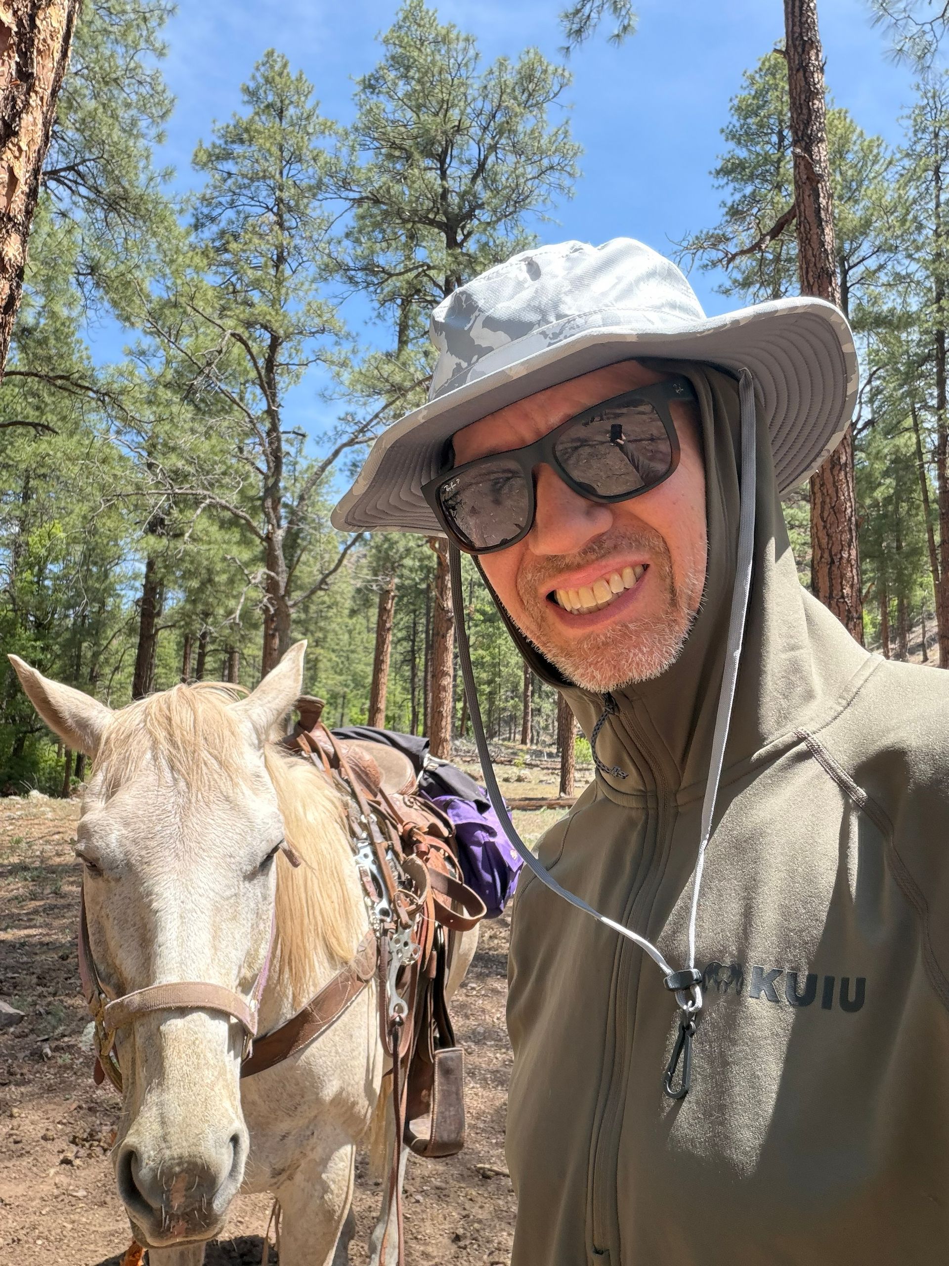 Man smiling beside a white horse in a sunny pine forest, wearing a hat and sunglasses.
