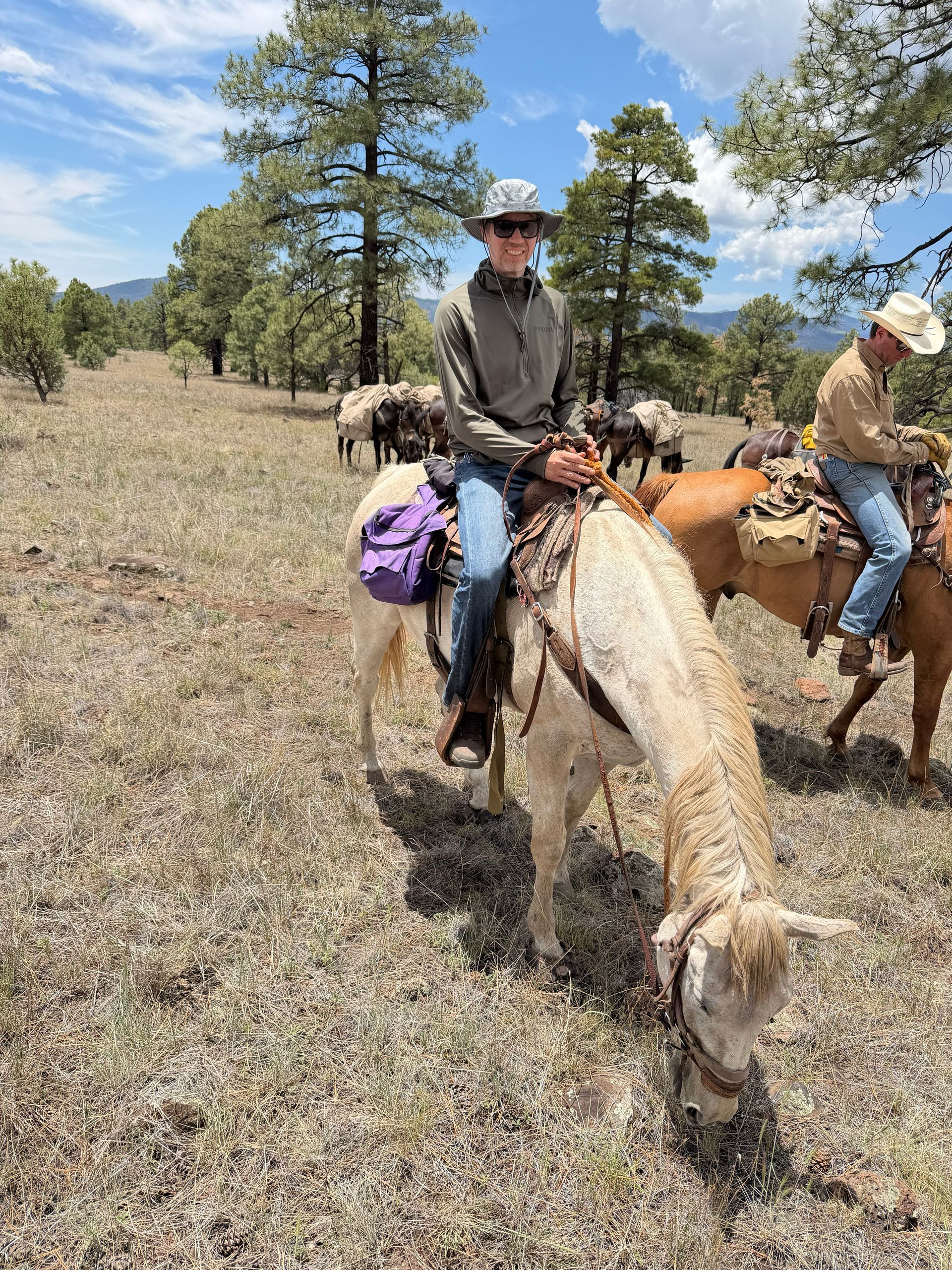 Two horseback riders in a dry, grassy field with pine trees and a blue sky.