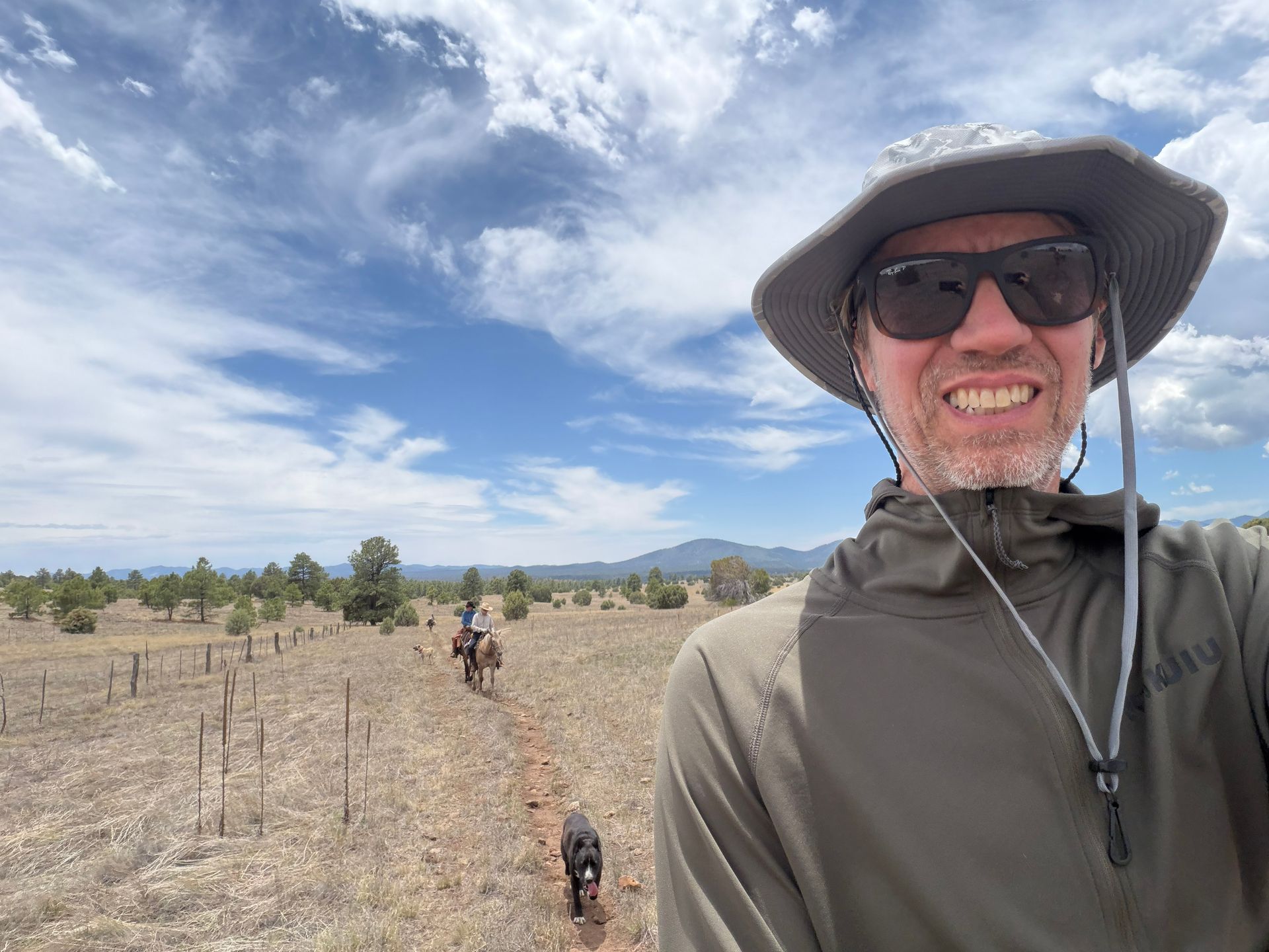 Smiling hiker in sun hat and sunglasses on a desert trail with two dogs under a cloudy sky