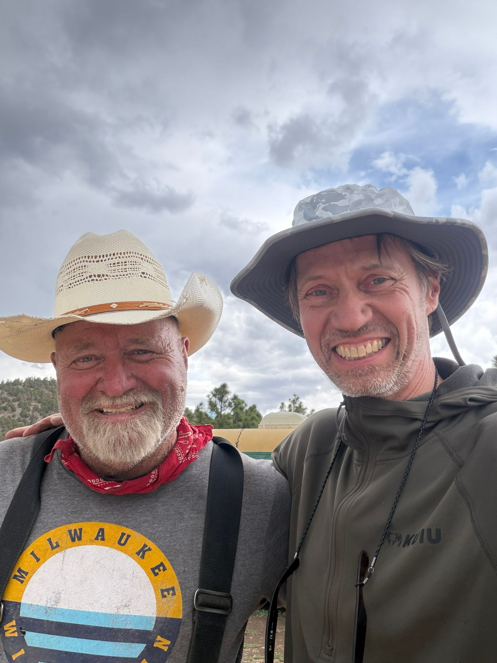 Two smiling hikers in sun hats pose outdoors under a cloudy sky.