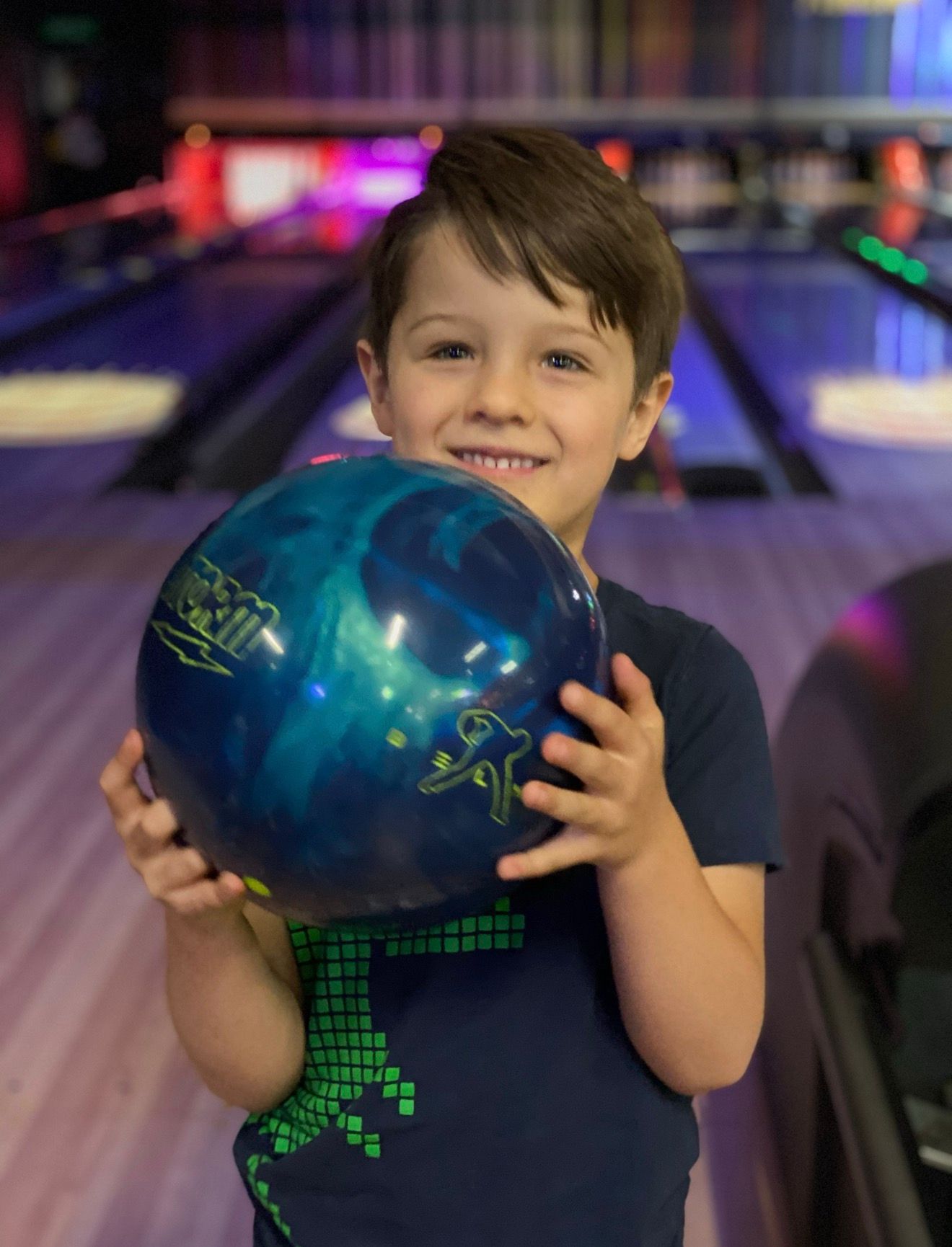 Child holding a blue bowling ball and smiling in a bowling alley lane area