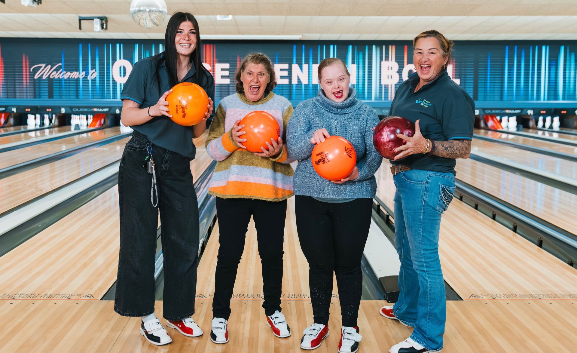 Four people posing with bowling balls at a bowling alley, with lanes behind them.