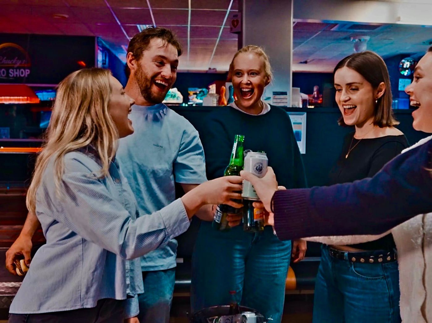 Friends clinking drinks and laughing at a lively indoor party with neon lighting.