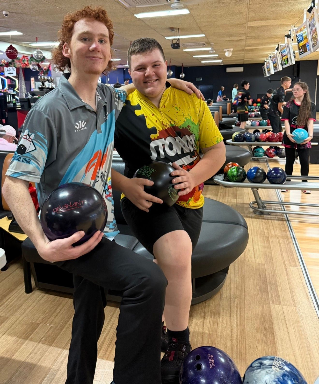 Two bowlers posing with bowling balls on a lane bench in a bowling alley, with lanes and pins behind them
