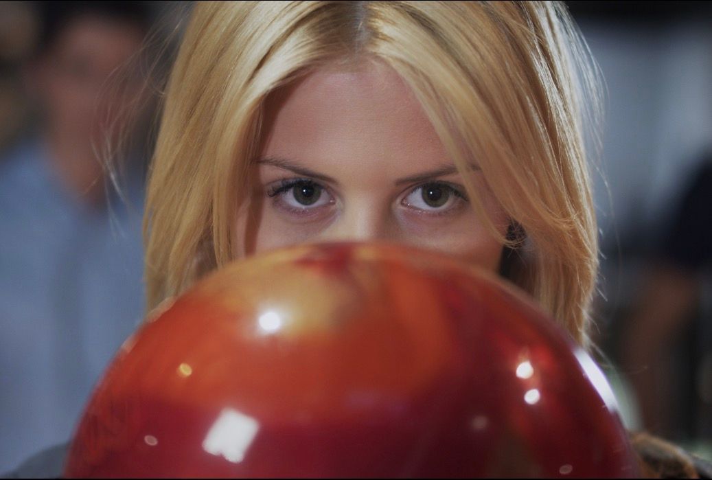 Blonde person staring over a glossy red bowling ball, focused in a bowling alley