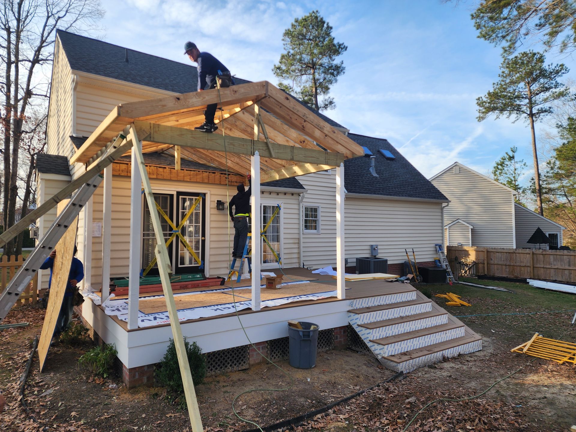 Construction workers building a wooden patio roof attached to a two-story house, on a sunny day.