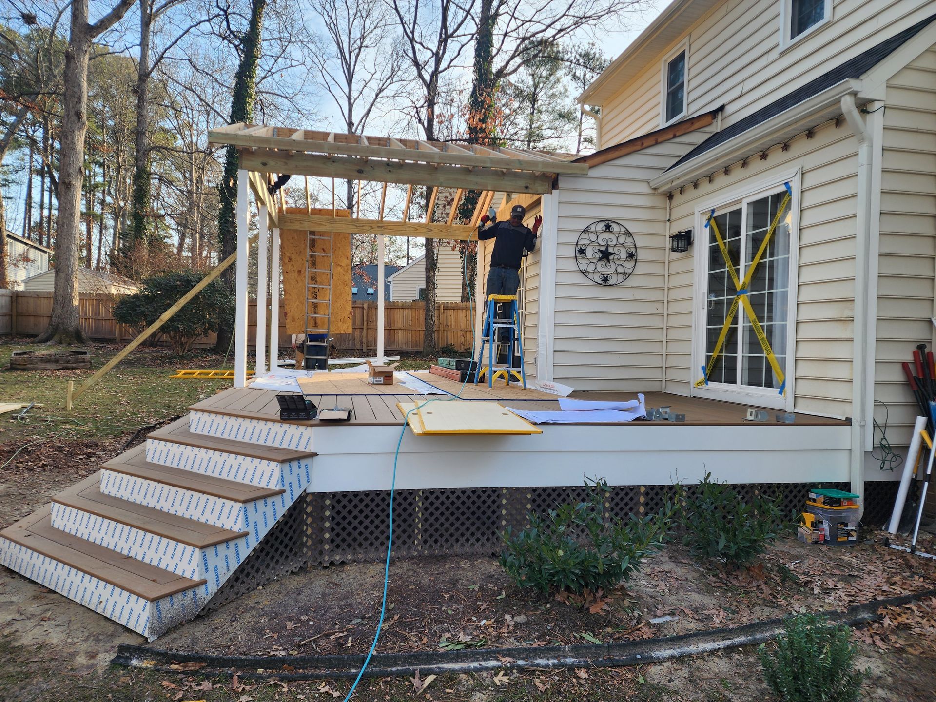 Deck construction: A person on a ladder works on a pergola attached to a house.