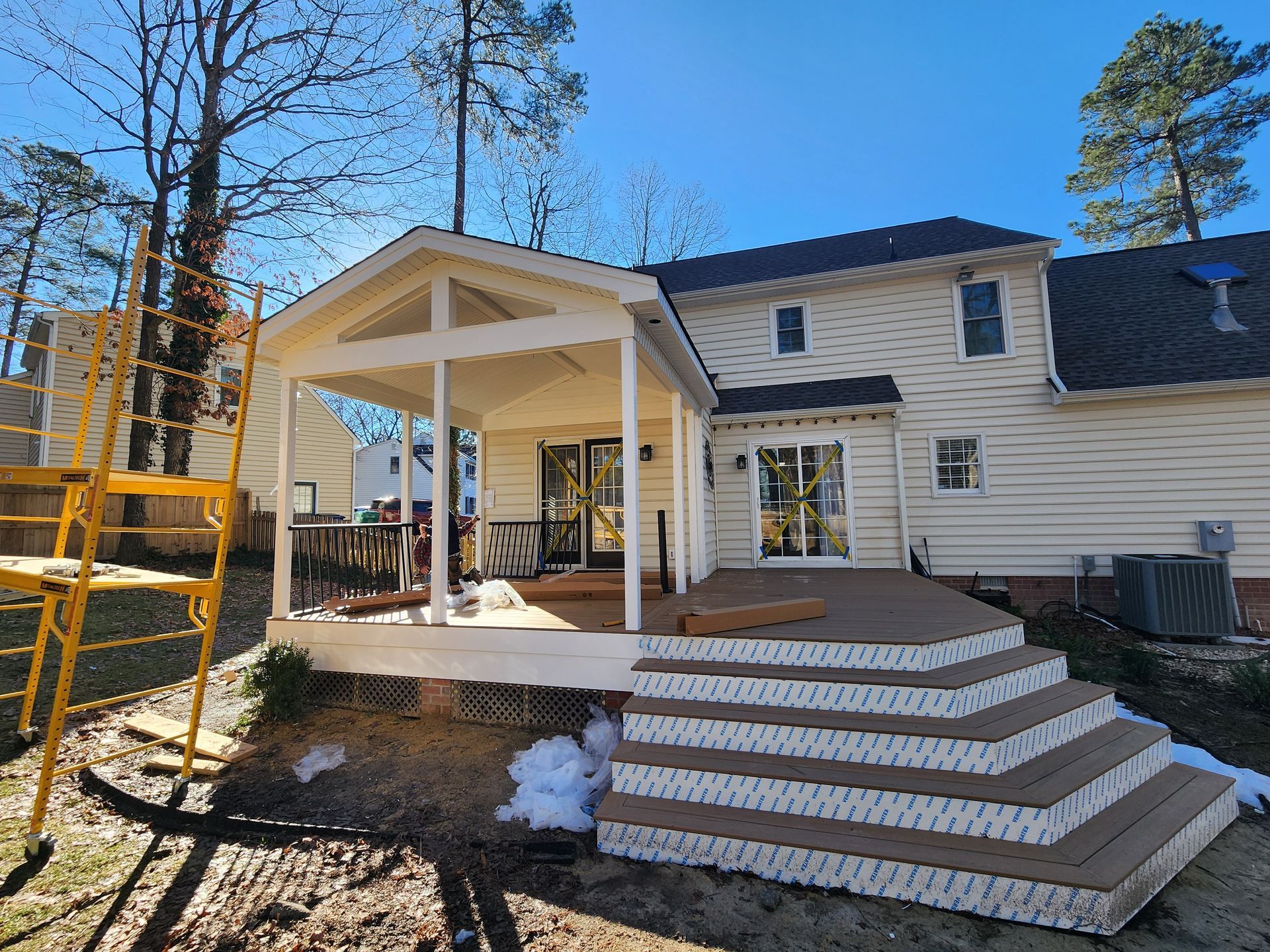 A back porch with a gabled roof extension, deck steps, and a house on a sunny day.