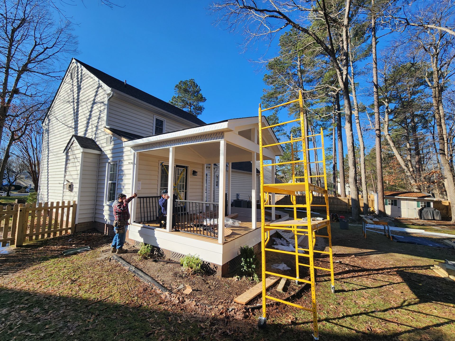 House with a porch under construction; yellow scaffolding; person standing nearby; sunny day.