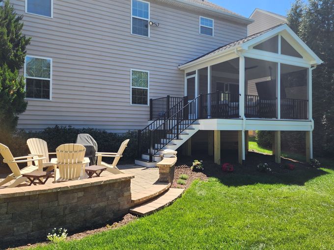Backyard with patio, raised screened porch, steps, and a two-story house. Green grass, wooden chairs, and a sunny day.