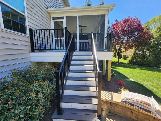 A screened porch with stairs, black railing, and gray deck. The lawn is green, with a red tree.