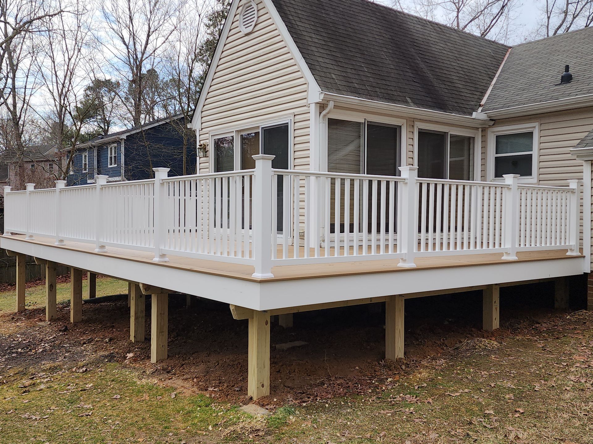 A white deck with railings attached to a beige house with a dark roof; green grass and trees in the background.