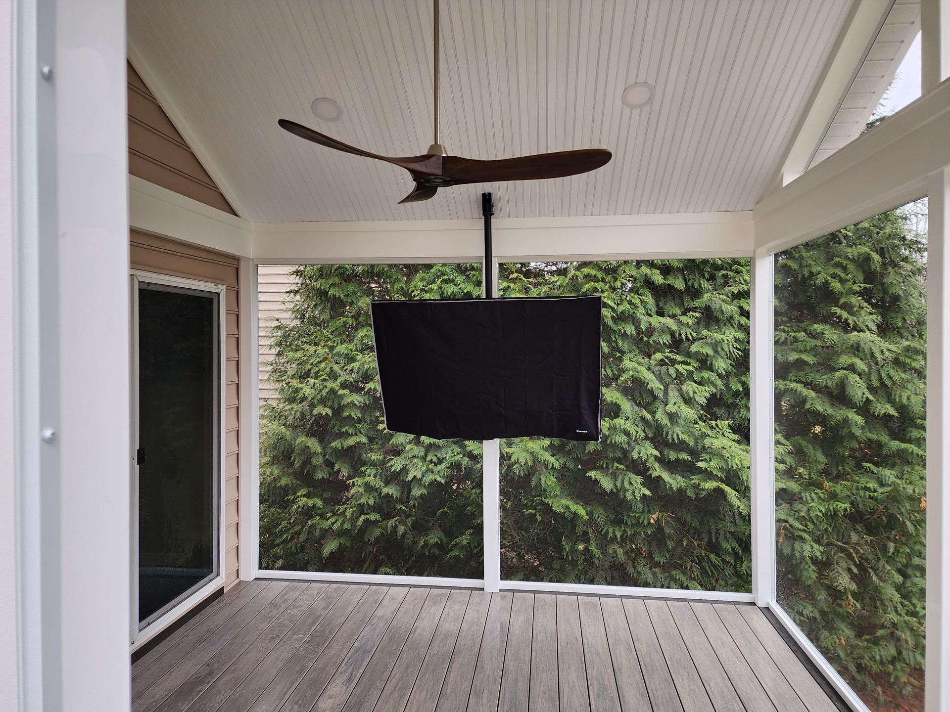 Covered porch with a TV mounted below a ceiling fan, overlooking a green yard.