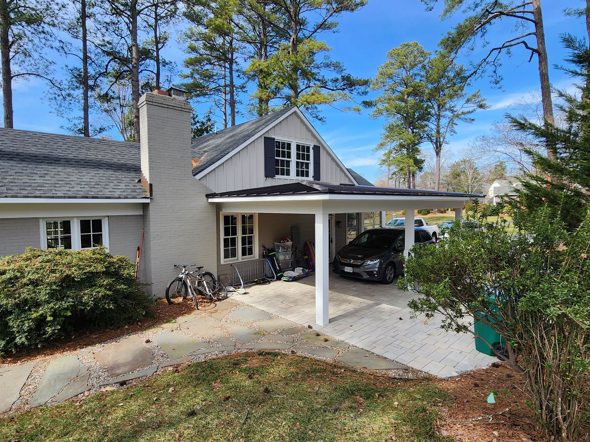 A house with a carport. A car and bikes are parked inside. Green lawn and trees surround the house.