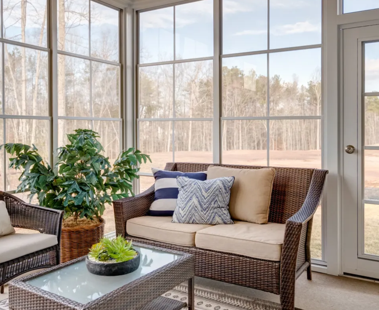 Sunroom with wicker furniture and throw pillows, overlooking a wooded area.